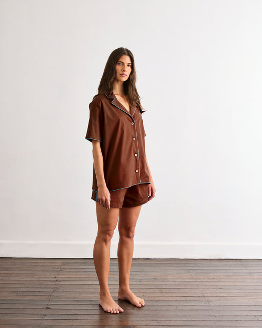 A woman with long brown hair stands barefoot on a wooden floor, wearing the Bed Threads Espresso & Sky Piped Short Sleeve Shirt—ideal for warmer nights—against a plain white wall.