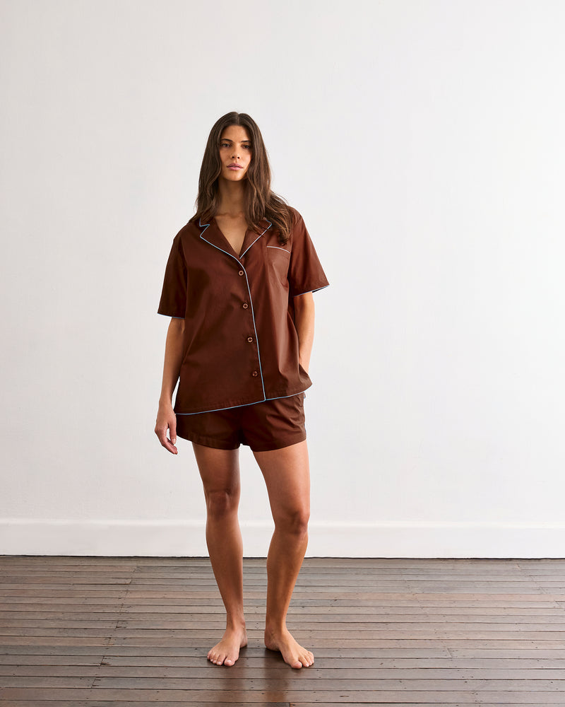 A woman stands barefoot on a wooden floor against a plain white wall, wearing Bed Threads’ Espresso & Sky Piped Shorts pajama set with white piping and breathable cotton fabric.
