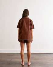 A barefoot person with long brown hair stands facing away on a wooden floor, wearing the Bed Threads Espresso & Sky Piped Short Sleeve Shirt—ideal for warmer nights—against a plain white wall. - view