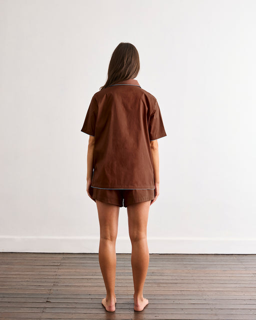 A barefoot person with long brown hair stands facing away on a wooden floor, wearing the Bed Threads Espresso & Sky Piped Short Sleeve Shirt—ideal for warmer nights—against a plain white wall.