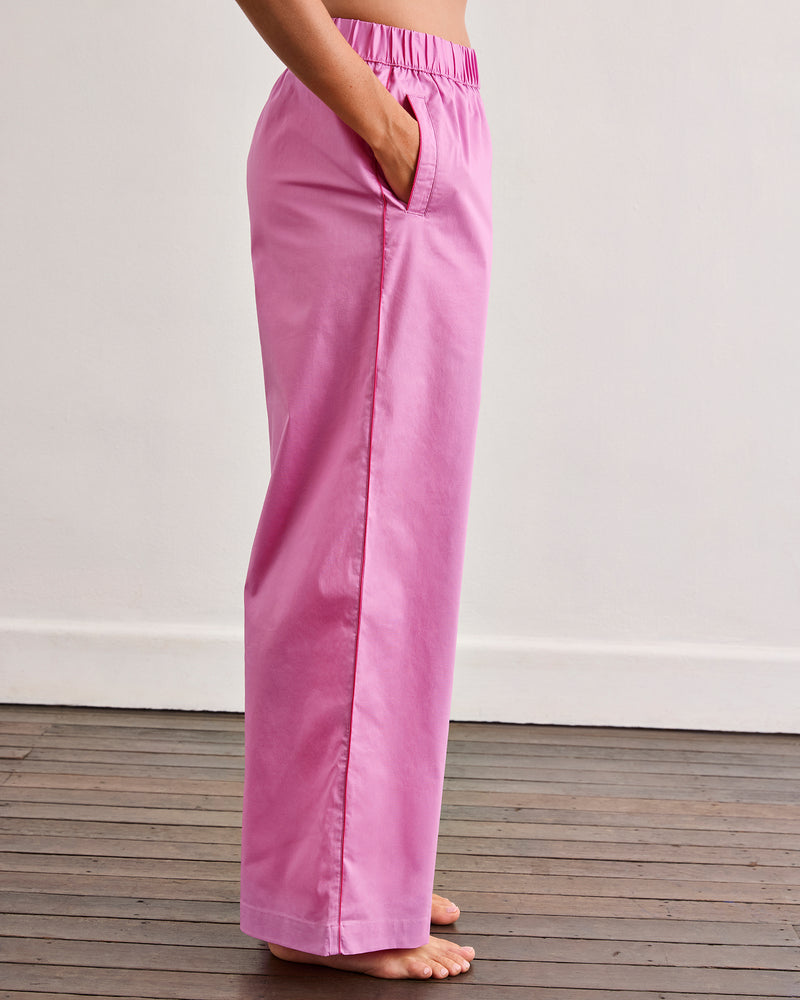 A person stands barefoot on a wooden floor, wearing Bed Threads’ Bubblegum & Berry Piped Pants in light pink cotton with an elasticated waist and side pockets, shown from the side to highlight their comfortable, relaxed fit.