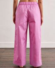A person stands barefoot on a wooden floor, shown from the waist down, wearing Bed Threads' Bubblegum & Berry Piped Pants—relaxed, high-waisted cotton pants with an elasticated waist. The background is a plain off-white wall. - view