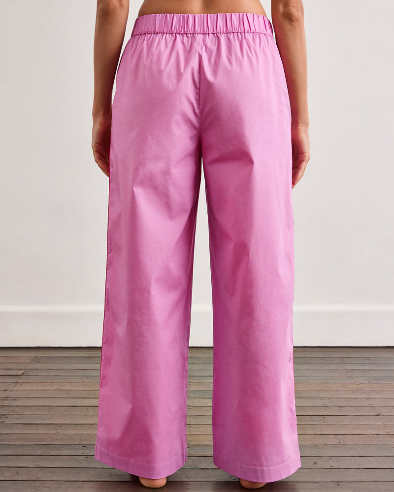 A person stands barefoot on a wooden floor, shown from the waist down, wearing Bed Threads' Bubblegum & Berry Piped Pants—relaxed, high-waisted cotton pants with an elasticated waist. The background is a plain off-white wall.