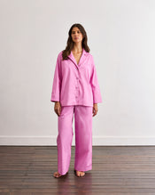 A person stands barefoot on a wooden floor, wearing Bed Threads' Bubblegum & Berry Piped Pants—relaxed, long-sleeved, button-up pink pajamas with an elasticated waist—in front of a plain white wall. - view