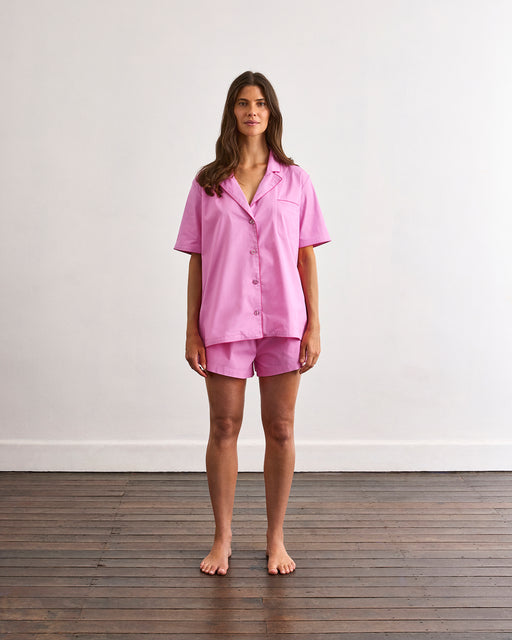 A woman stands barefoot on a wooden floor against a white wall, wearing the Bed Threads Bubblegum & Berry Piped Short Sleep Set—featuring matching pink cotton piped shorts and a short sleeve shirt.