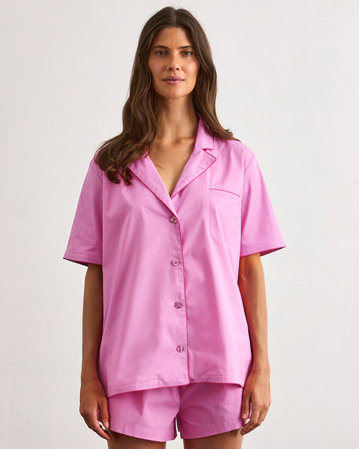 A person with long brown hair stands against a light background, wearing the Bubblegum & Berry Piped Short Sleeve Shirt by Bed Threads—a matching pink cotton pajama top and shorts set.