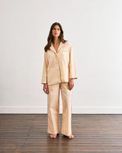 A woman with long brown hair stands barefoot on a wooden floor, wearing the Bed Threads Vanilla & Espresso Piped Long Sleeve Shirt, paired with wide-leg pants, against a plain white wall. - view