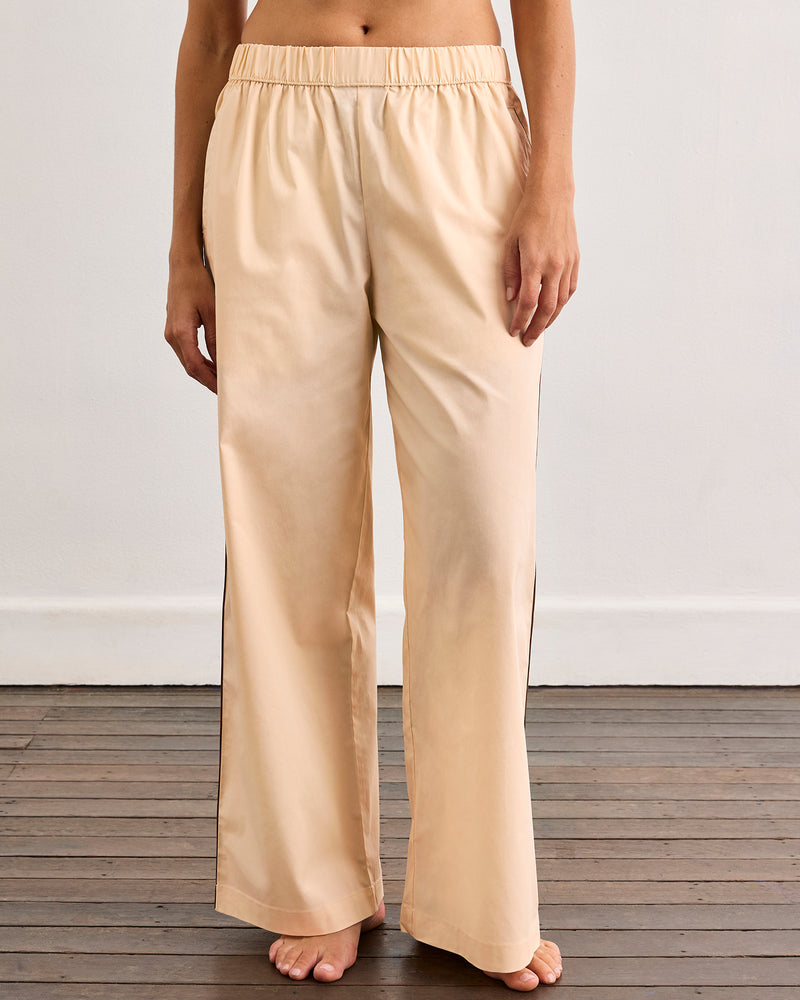 A person stands barefoot on a wooden floor, wearing loose, light beige pajama pants from the Vanilla & Espresso Piped Long Sleep Set by Bed Threads. The photo is cropped to show only the lower half of their body.