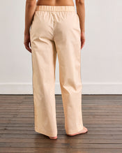 A person stands barefoot on a wooden floor, modeling Bed Threads’ Vanilla & Espresso Piped Pants—relaxed cotton trousers with a comfy elastic waist—shown from the back against a plain white wall. - view