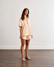 A woman stands barefoot on a wooden floor, wearing the Bed Threads Vanilla & Espresso Piped Short Sleeve Shirt and matching shorts, made of lightweight cotton. She poses against a plain white wall, looking slightly to the side. - view