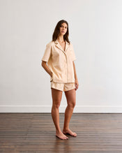 A woman stands barefoot on a wooden floor against a plain white wall, wearing Bed Threads' Vanilla & Espresso Piped Shorts—a short-sleeve, button-up beige pajama shirt and matching breathable shorts. - view