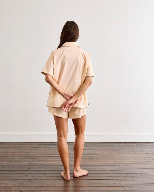 A woman with long, loose hair stands barefoot on a wooden floor facing a white wall, hands behind her back. She wears the Bed Threads Vanilla & Espresso Piped Short Sleeve Shirt and matching shorts, creating a relaxed look.