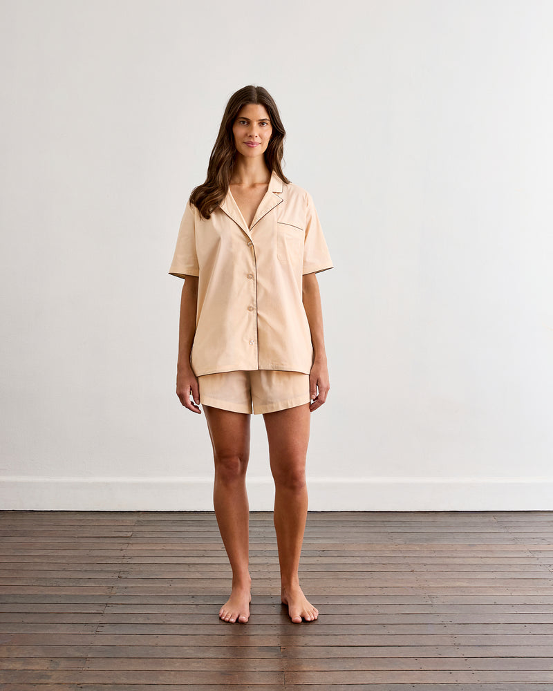A woman stands barefoot on a wooden floor in front of a white wall, wearing the Bed Threads Vanilla & Espresso Piped Short Sleep Set—a lightweight cotton short-sleeve shirt and piped shorts.
