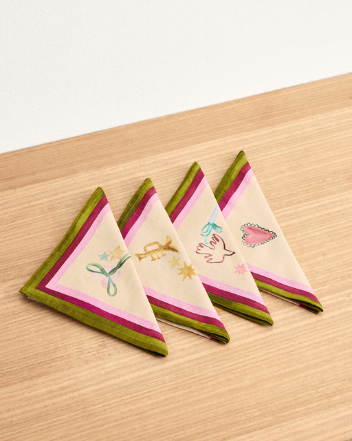 Four La Fête 100% French Flax Linen Napkins by Bed Threads are folded into triangles on light wood. Each festive napkin features green and pink borders with whimsical motifs like a hand, heart, ribbon, and stars. Set of four.
