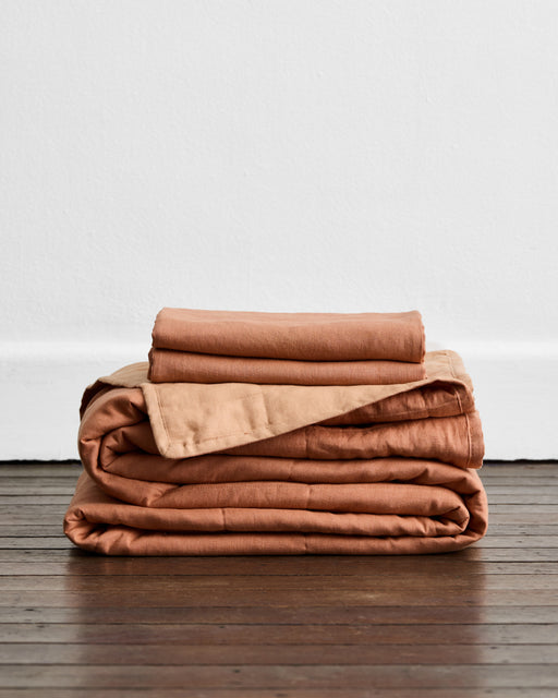 A neatly folded Bed Threads Terracotta & Hazelnut 100% French Flax Linen Quilt Set rests on a wooden floor against a plain white wall.