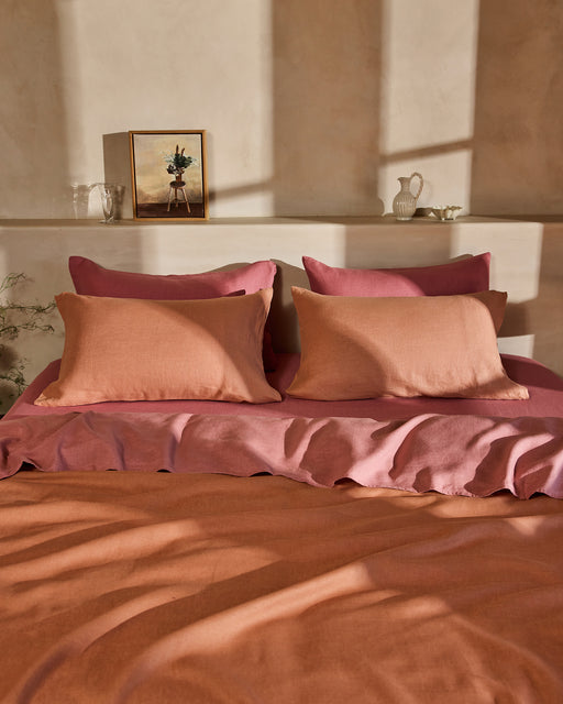 A bed dressed in the Bed Threads Hazelnut & Pink Clay 100% French Flax Linen Bedding Bundle sits against a beige wall, with a shelf above holding decor like a framed painting, glass, white vase, and ceramic pitcher. Shadows fall across the scene.