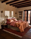 A cozy bedroom features a double bed dressed in Bed Threads' Hazelnut & Pink Clay 100% French Flax Linen Bedding Bundle, set atop a red rug, with wood beams, beige walls, built-in shelves, wall sconces, and sunlit glass doors.