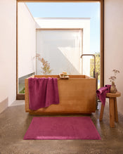 A modern bathroom with a wooden bathtub, magenta towels and bath mat, a small wooden stool with a vase, and large windows letting in natural light. Minimalist decor and concrete floor create a serene atmosphere. - view