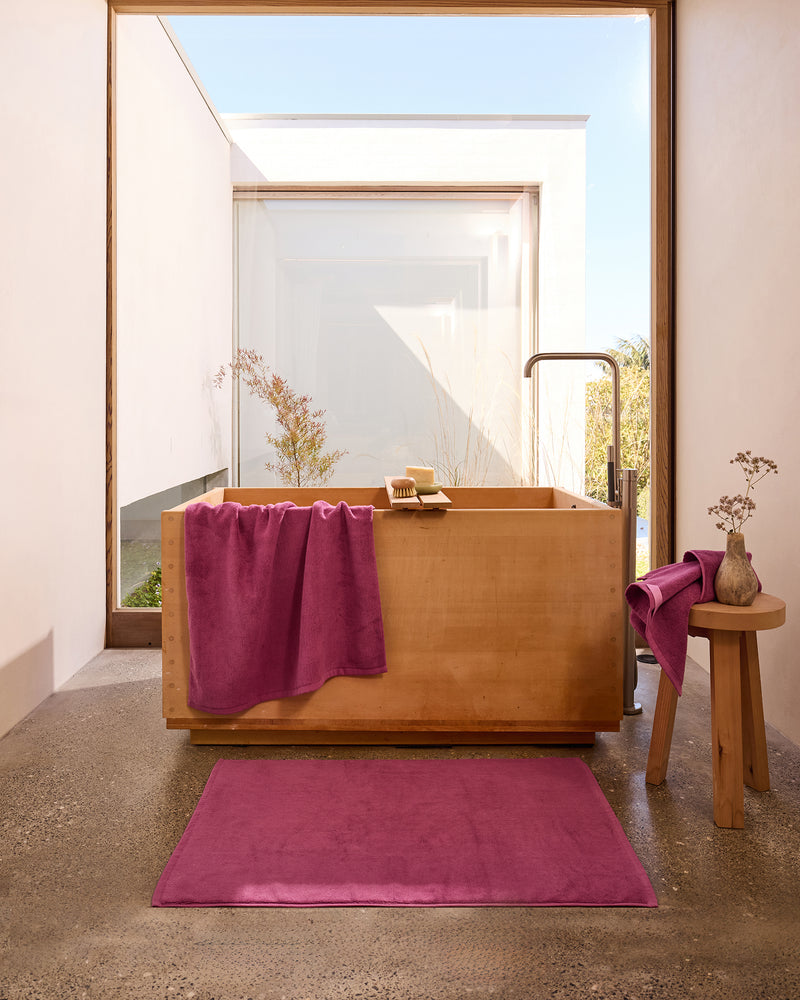 A modern bathroom with a wooden bathtub, magenta towels and bath mat, a small wooden stool with a vase, and large windows letting in natural light. Minimalist decor and concrete floor create a serene atmosphere.