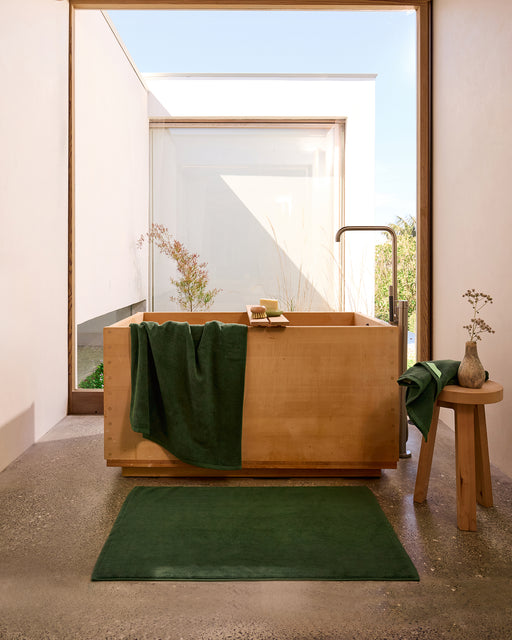 A modern bathroom features a wooden soaking tub, Bed Threads Moss Cotton Terry Bath Towels, a matching green bath mat, sunlight streaming through a large window, and a vase of flowers on a wooden stool.