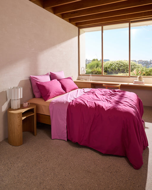 A minimalist, cozy bedroom features a bed with bright pink and light purple bedding, accented by Bed Threads' Bubblegum 100% Organic Cotton European Pillowcases (Set of Two), wooden nightstands, and large windows overlooking greenery.
