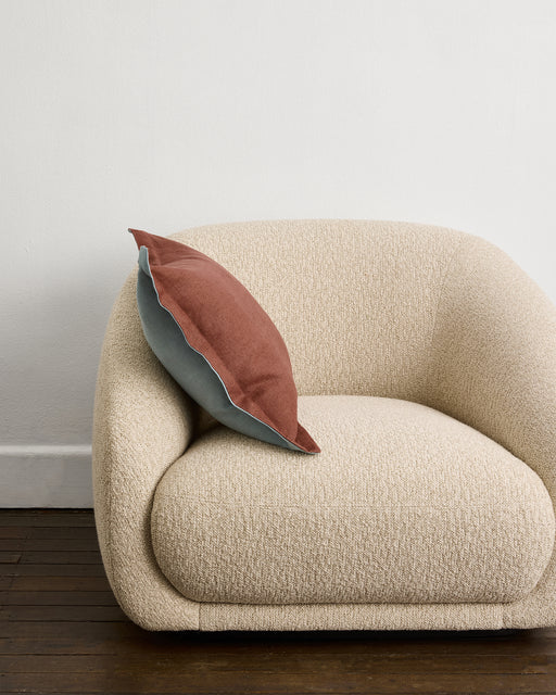 A modern, cream-toned textured armchair sits on a dark wooden floor. A Bed Threads Cacao & Lagoon 100% French Flax Linen Cushion Cover rests on the chair, with its rust and light blue sides visible. A plain white wall is in the background.