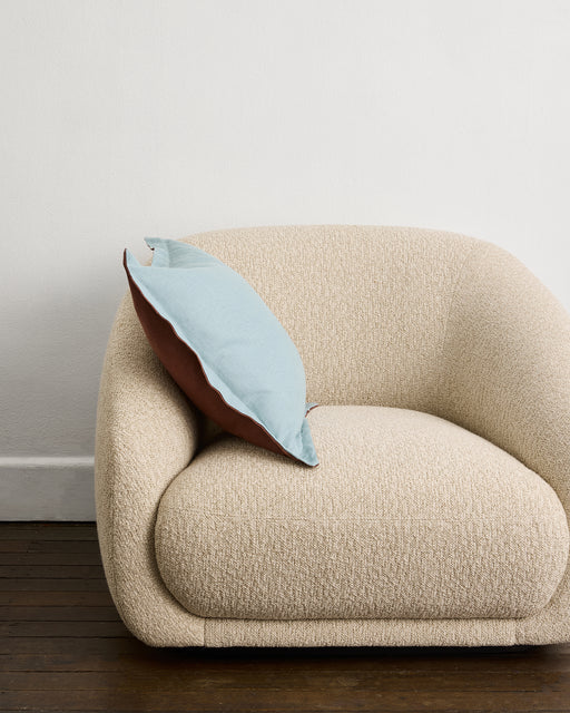 A beige, modern boucle armchair sits on a dark wooden floor. Resting against one arm is the Cacao & Lagoon 100% French Flax Linen Cushion Cover by Bed Threads. The background features a plain white wall.