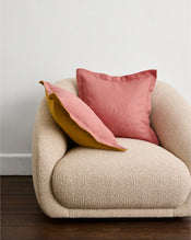 A plush cream armchair with two square cushion covers—one Pink Clay & Turmeric 100% French Flax Linen Cushion Cover by Bed Threads—stands on wooden flooring against a plain white wall. - view