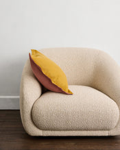 A cream-colored, rounded armchair featuring a Bed Threads Pink Clay & Turmeric 100% French Flax Linen Cushion Cover rests on a dark wooden floor against a plain white wall. - view