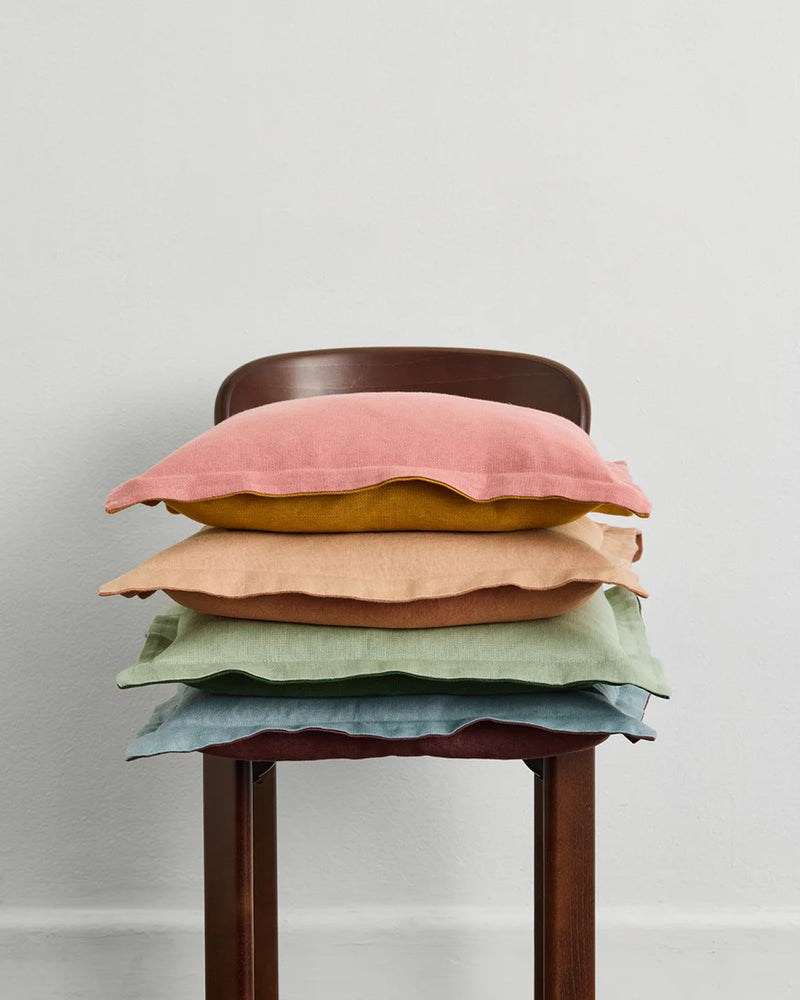 A stack of Bed Threads Pink Clay & Turmeric 100% French Flax Linen Cushion Covers brightens a wooden chair set against a plain white wall.