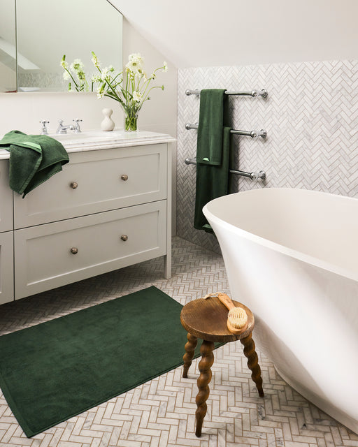 A modern bathroom with a white freestanding tub, marble-topped vanity, Bed Threads Moss 100% Cotton Terry Bath Towels (Set of Two), matching bath mat, stool with hairbrush, herringbone tile throughout, and a vase of flowers for a complete look.