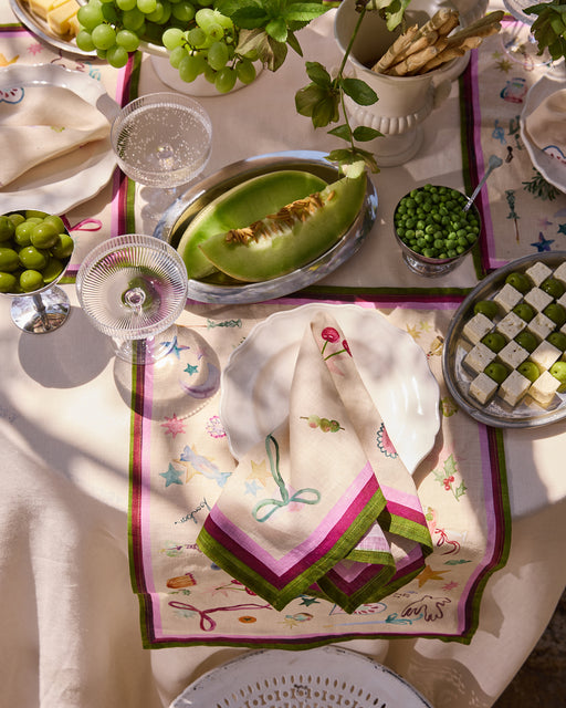 A sunlit table set for a meal with grapes, melon slices, green peas, cheese cubes, breadsticks, and Bed Threads’ La Fête 100% French Flax Linen Napkins (Set of Four) on white plates, amid glassware and fresh produce for a festive touch.