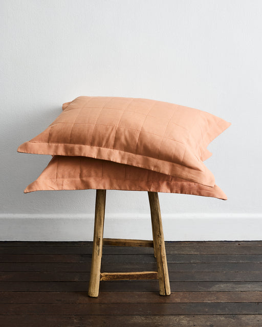 Two Terracotta Quilted Pillowcases by Bed Threads, featuring a subtle grid pattern and made from French flax linen, are stacked on a rustic wooden stool against a white wall and dark wood floor.