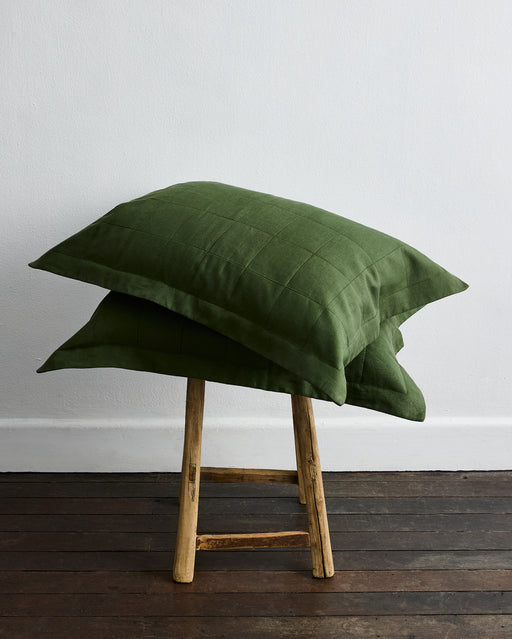 Two Olive Quilted Pillowcases by Bed Threads are stacked on a small wooden stool, set against a white wall and dark wood floor.