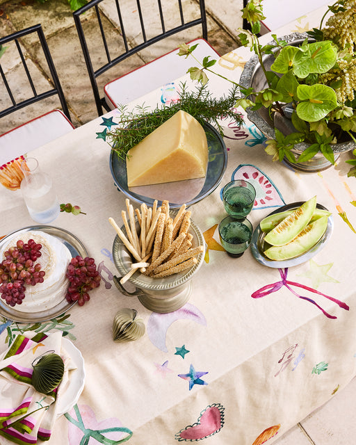 A vibrant outdoor table is set with the limited-edition La Fête 100% French Flax Linen Tablecloth by Bed Threads, featuring cheese, breadsticks, melon slices, grapes, green glasses, a glass of water, potted plants, and metal chairs.