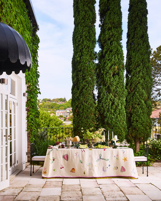 A patio dining table with a colorful, limited-edition La Fête 100% French Flax Linen Tablecloth by Bed Threads is set amid cypress trees beside a white house with French doors and ivy, overlooking a lush, hilly neighborhood.