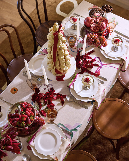 A festive dining table features white plates, candles, a centerpiece tree of white chocolates, grapes, berries, a halved pomegranate, and Bed Threads’ La Fête 100% French Flax Linen Napkins (Set of Four), surrounded by four wooden chairs.