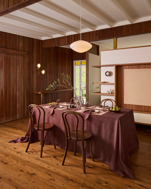 A cozy dining room features wood-paneled walls and floors, a mauve-covered table with four wooden chairs, Bed Threads’ Hazelnut 100% French Flax Linen Placemats (Set of Four), simple tableware, pendant lighting, minimalist art, and tall branches in a vase.