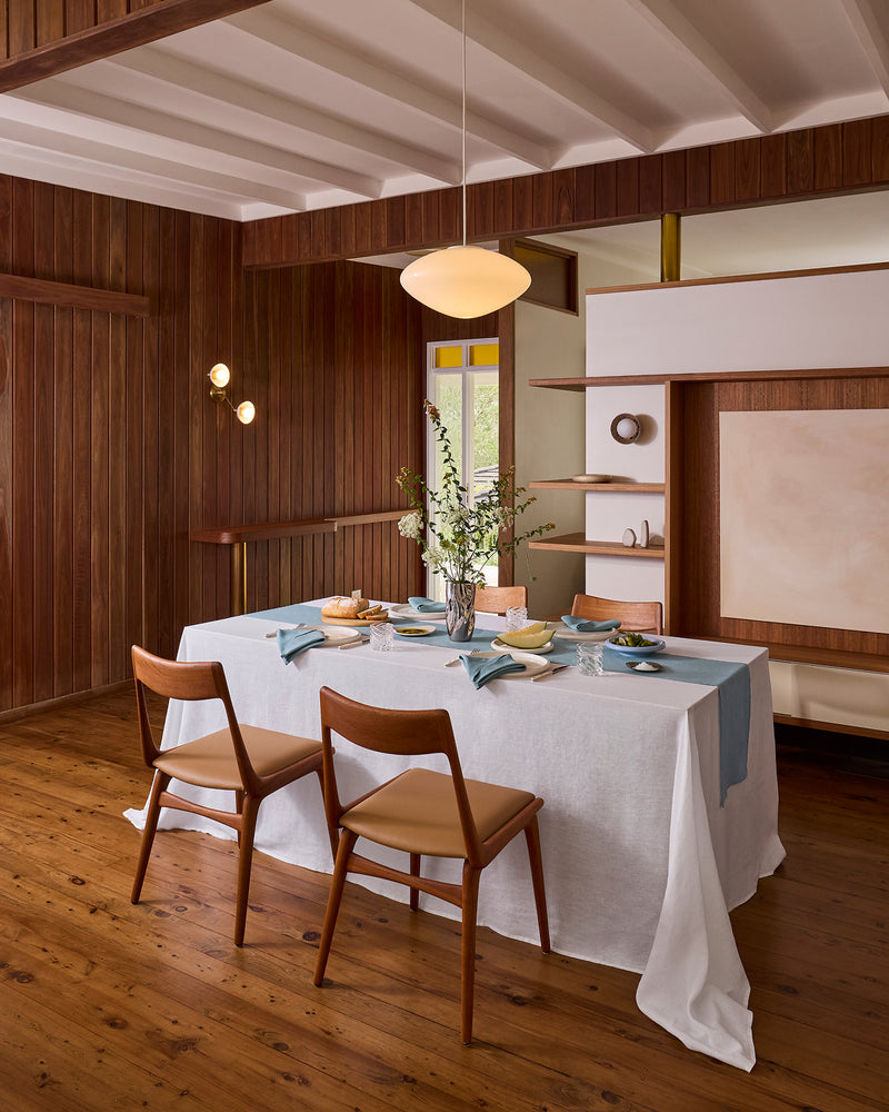 A dining room with wood-paneled walls and floor features a Bed Threads White & Lagoon 100% French Flax Linen Table Bundle on the table, set for a meal with dishes and glassware. Two wooden chairs are placed at the table.
