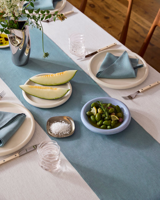 A dining table features the Lagoon 100% French Flax Linen Table Runner by Bed Threads, white plates with blue napkins, clear glasses, silverware, sliced melon, green olives with lemon, and a small dish of salt.