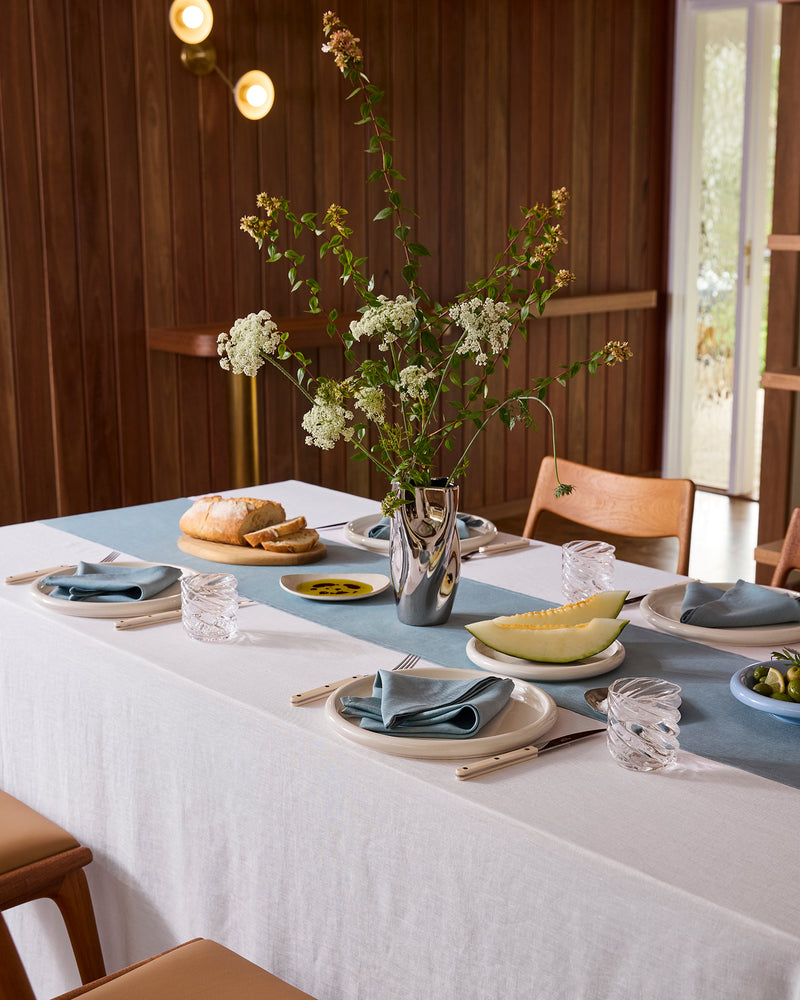 The White & Lagoon 100% French Flax Linen Table Bundle from Bed Threads adds elegance with a white tablecloth, classic napkins, blue glassware, plates, bread, melon slices, olives, and a silver vase. Wood accents create a warm modern setting.