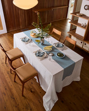 A dining table with a white tablecloth and the Bed Threads Lagoon 100% French Flax Linen Table Runner is set with plates, glasses, napkins, cutlery, a vase of flowers, and fruit bowls. Four wooden chairs are arranged on the wooden floor. - view