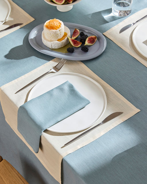 A dining table is set with a white plate, silverware, and a folded Lagoon 100% French Flax Linen Napkin by Bed Threads on a cream and blue tablecloth. Nearby is a plate of dessert, figs, and blackberries.
