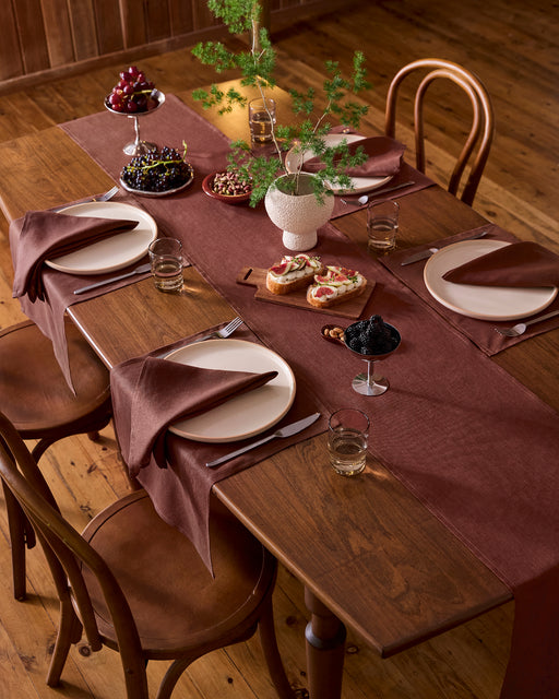 A wooden dining table set for three features brown napkins and runner, Bed Threads’ Cacao 100% French Flax Linen Placemats, white plates, glasses, a potted plant centerpiece, assorted desserts, fruit, and open-faced sandwiches on a wooden floor.