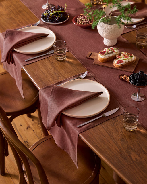 A wooden dining table set for two features Bed Threads' Cacao 100% French Flax Linen Placemats, white plates, brown napkins, glasses of water, cutlery, snacks, fruit bowls, appetizers, and a potted plant.
