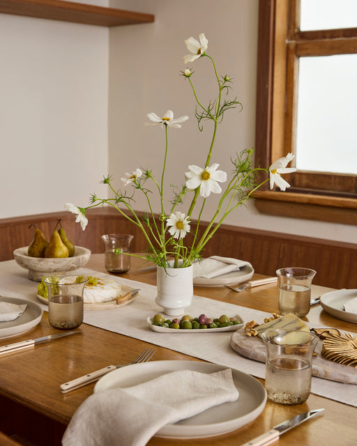 A wooden dining table set for four features white plates, linen napkins, water glasses, a vase of white flowers, pears, cheese, and olives atop a Bed Threads Oatmeal 100% French Flax Linen Table Runner as sunlight streams in.