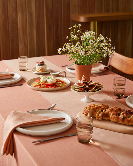 A dining table features the Bed Threads Terracotta 100% French Flax Linen Table Runner, set with plates, glasses, napkins, bread, figs, tomato salad, dessert, and an elegant daisy centerpiece for a charming touch.