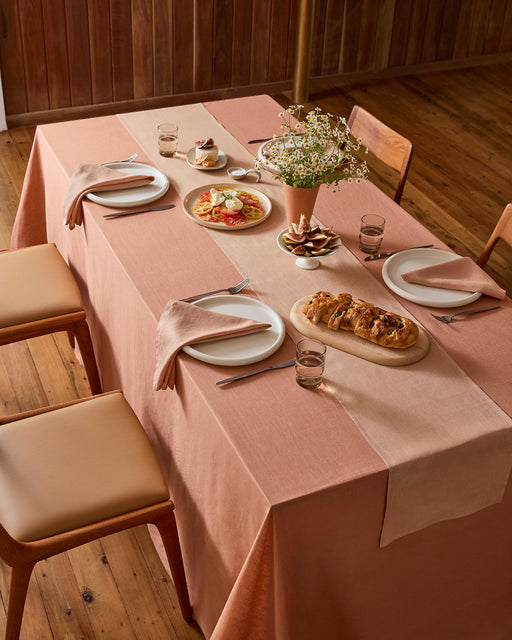 A wooden dining table for four is set with a pink tablecloth, Bed Threads Terracotta 100% French Flax Linen Table Runner, neutral napkins, plates of pastries and appetizers, glasses of water, and a vase of daisies.