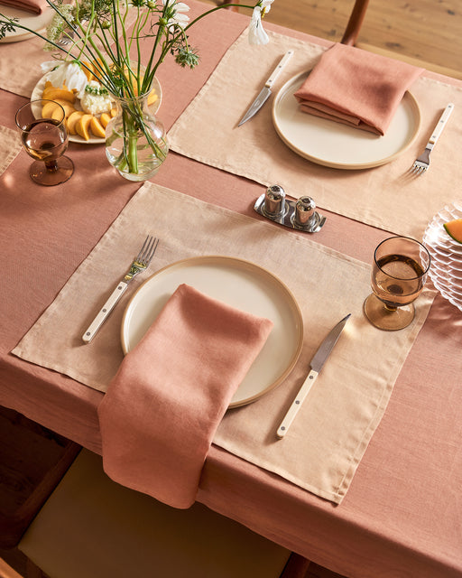 A table set for two with the Hazelnut & Terracotta 100% French Flax Linen Table Bundle by Bed Threads, white plates, pink napkins, silver cutlery, amber glassware, flowers, lemons, and salt and pepper shakers creates a cozy dining scene.