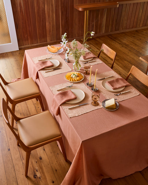 A white plate with crossed fork and knife rests on a Bed Threads Terracotta 100% French Flax Linen Placemat atop a rustic wooden table, with a spoon to the right and a clear glass above for stylish table protection.